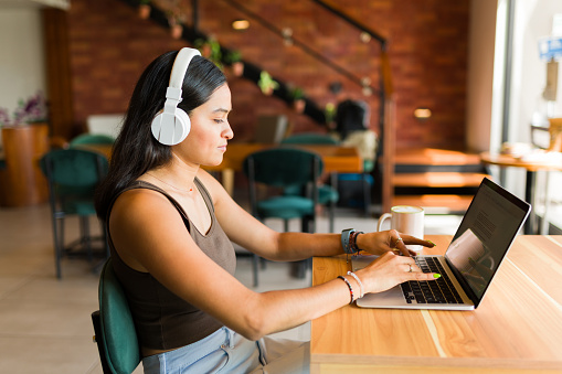 Hispanic young woman student with headphones listening to music and doing homework and working at the coffee shop