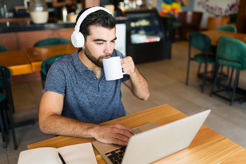 Attractive young man doing freelance or remote work on the laptop using wifi while drinking coffee at the cafe