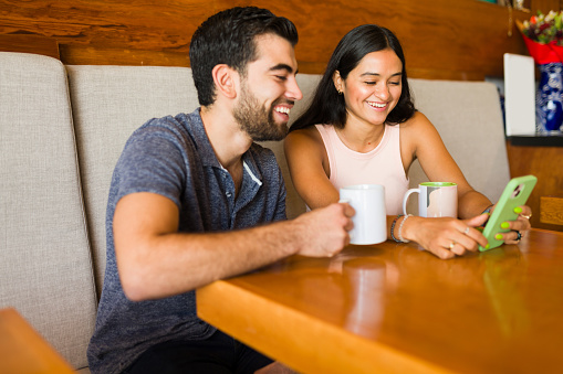 Smiling attractive man and woman having a date at a coffee shop and texting on the smartphone while looking happy