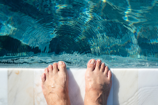Close-up at human barefeet which is standing on the edge of swimming pool with cleary turquoise water shade. Recreation activity for vacation concept.