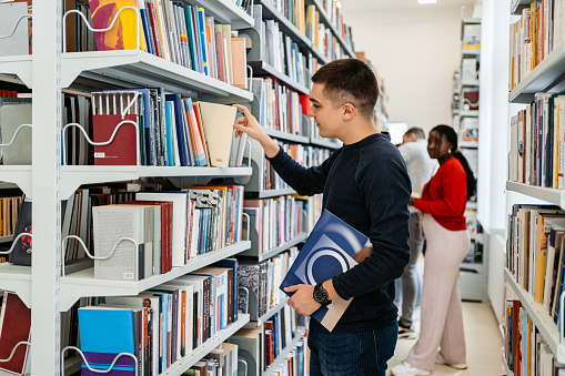 Young male student picking up books in a student library in college.
