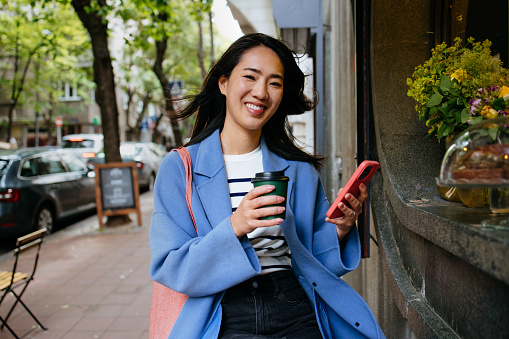 A portrait of a smiling Japanese woman in the city, using her smartphone and having a coffee.