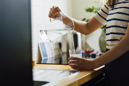 An unrecognizable woman pouring a glass of water from a pitcher in the kitchen.