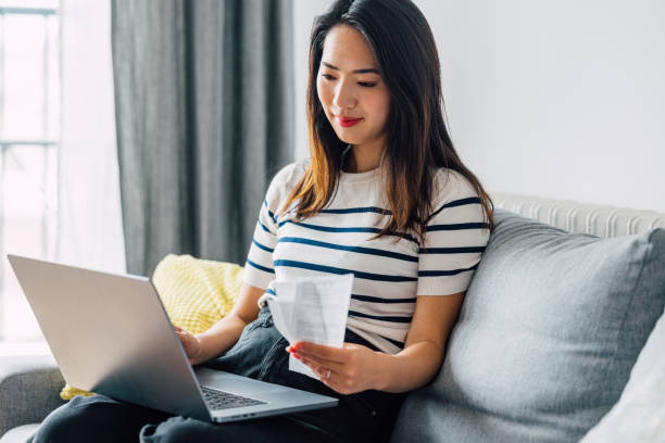 istock Young Asian Woman Analyzing House Bills while Sitting on the Sofa with a Computer on her Lap 1546277550