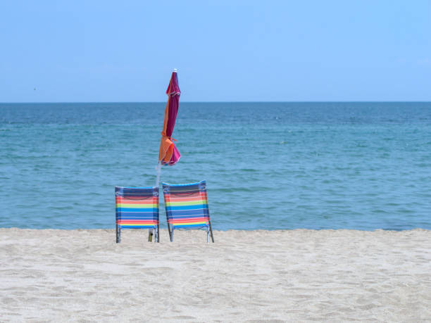istock Two empty beach chairs with a rainbow design and a closed umbrella stand on a sandy beach by the sea 1546665064