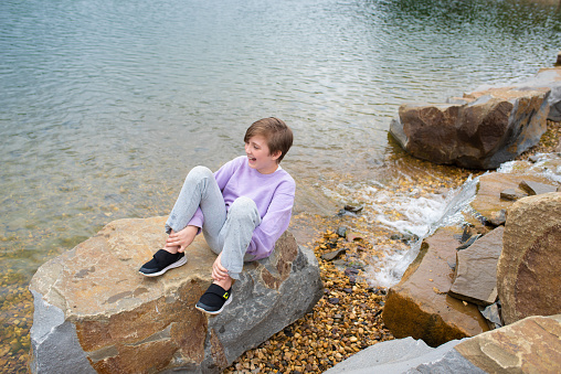 the child smiles and laughs, sits on a large stone in front of a small waterfall near the lake in cloudy weather. happy emotional boy resting in nature in summer.