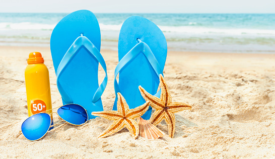 STILL LIFE COMPOSED OF FLIP FLOP, SUNGLASSES, FACTOR 50+ SUNSCREEN LOTION AND TWO STARFISH ON THE SAND OF THE BEACH. SKIN SOLAR PROTECTION ON VACATIONS.