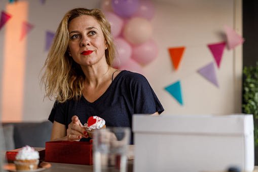 Copy space shot of happy mid adult woman sitting at table and packing Woman cute Valentine's day themed cupcake in a red gift box. She is looking away, smiling and contemplating.