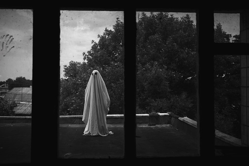 Ghost in a sheet on the roof of an old abandoned house view through the window, spooky Halloween concept, copy space