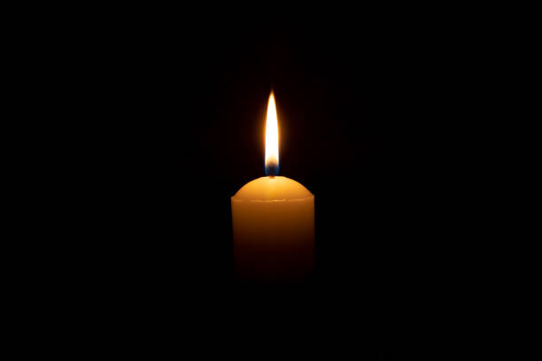istock Single burning candle flame or light glowing on a big yellow candle on black or dark background on table in church for Christmas, funeral or memorial service with copy space. 1547293282