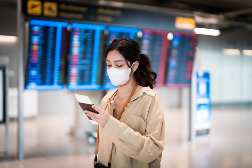 Young asian woman wearing protective virus face mask with passport and boarding pass as a hand in international airport looking at the flight information board, checking her flight