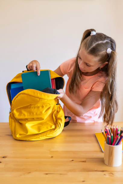 Blonde Elementary School Girl With Pigtails Puts Office Supplies Into A Backpack.