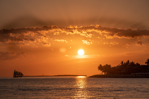 orange sky, sun, sailing boat, reflections in water, waves sea, tropical trees, clouds, cloud shadows