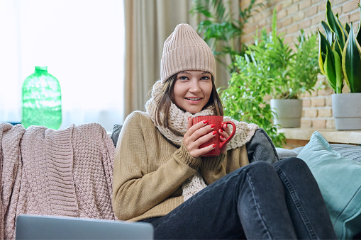 Cold season autumn winter, young woman in cozy woolen sweater hat knitted scarf sitting at home on sofa, basking with mug of hot drink tea. Winter cold holidays, relax, comfort, coziness, lifestyle