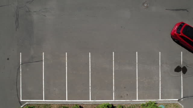 A red car pulls out of a parking space and drives away, Aerial shot. Empty parking lot. Smooth white markings in the parking lot. The car is parked, top view.