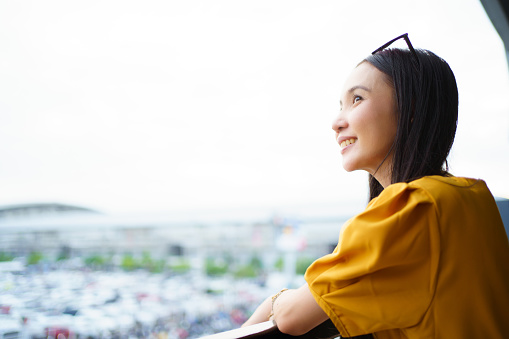 Beautiful happy Asian young woman in a beautiful yellow dress standing on the building balcony and enjoy looking outside at the sky and city view with copyspace. Simply and carefree lifestyles concept.
