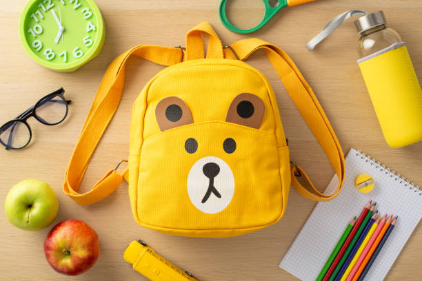 istock Primary education concept. Top-view image of assorted supplies, notepad, color pencils, sharpener, glasses, clock, toy, magnifier, playful bear-shaped backpack, water bottle, and apples on wooden desk 1547641753