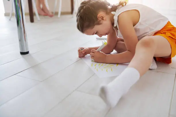 Caucasian cute baby girl with two pigtails, sitting on the floor, focused on drawing beautiful picture with watercolor felt-tip-pens at home. People. Kids. Childhood. Lifestyle. Hobby. Painting. Art