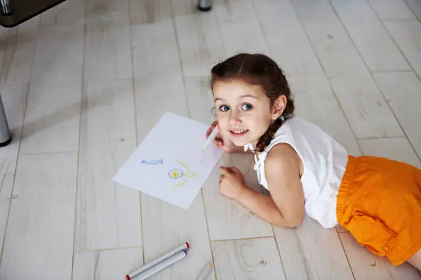 Overhead view Caucasian beautiful lovely preschooler girl drawing picture on white paper sheet with watercolor felt-tip-pens, lying on the floor, smiling cutely looking at camera. Kids. Hobby. Leisure