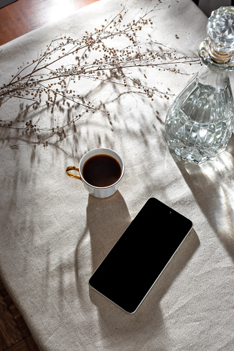 Aesthetic minimalist business morning scene, mobile phone, cup off coffee, glass vase and aesthetic sunlight shadows on a neutral beige table cloth