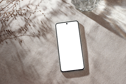 Mobile phone with blank screen mock up on a neutral beige linen background with aesthetic floral sunlight shadows, minimalist business brand, social media blog template, copy space