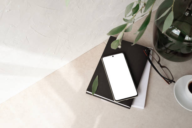 istock Mobile phone with empty screen mockup, notebook, glasses, vase with foliage branch, empty neutral beige background with aesthetic sunlight shadow, minimalist business brand, social media blog template 1547976057