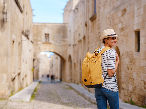 summer trip to Rhodes island Greece. Young Asian woman in striped tshirt and hat walks Street of Knights of Fortifications castle. female traveler visiting southern Europe. Unesco world heritage site