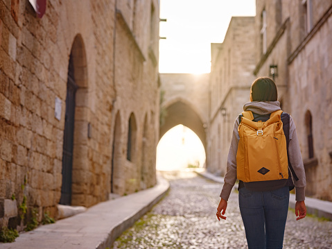 spring or fall trip to Rhodes island, Greece. Young Asian woman walks Street of Knights of Fortifications castle over sunset time. female traveler visiting southern Europe. Unesco world heritage site.