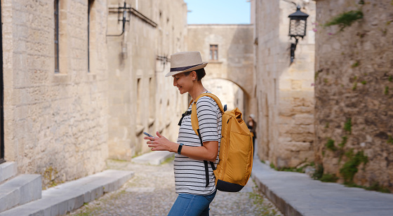 summer trip to Rhodes island Greece. Young Asian woman make photo on smartphone walks Street of Knights of Fortifications castle. female traveler visiting southern Europe. Unesco world heritage site