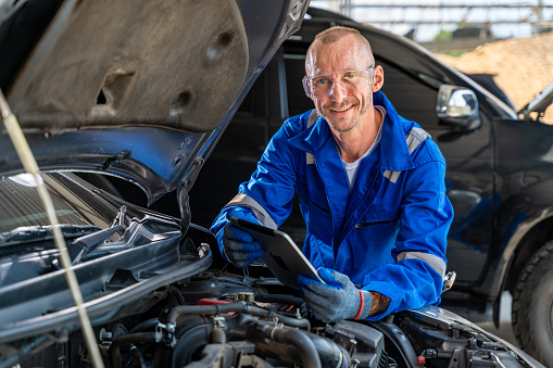 Happy car mechanic holding a computer tabet and smiling at camera while working at a car's engine
