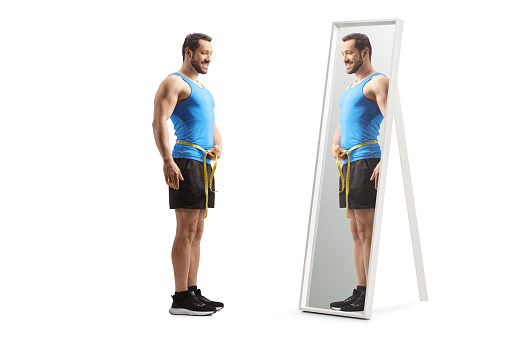 Full length shot of a fit man in sportswear measuring waist and looking at a mirror isolated on white background