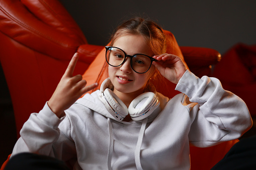 Attractive stylish little girl child wearing a casual sweatshirt and headphones, showing rock n roll sign and touching her glasses sitting in chair and looking in camera. Childhood lifestyle concept