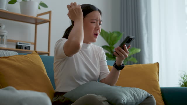 Asian woman feeling sadness reading or watching something at smartphone, sitting on sofa in living room at home.