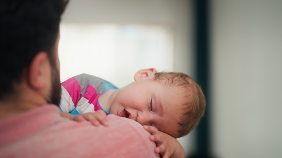 A father is carrying his small baby boy and comforting him in the living room at home.