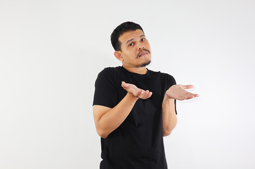 Young adult asian man raised arms and looking at camera with doubtful face. studio shot, isolated on light grey background.