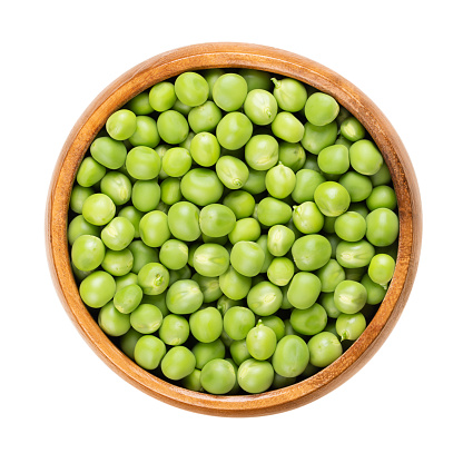 Fresh green peas, in a wooden bowl. Raw, small spherical seeds of the pod fruit Pisum sativum of greenish and yellowish color, mostly used for soups and as side dish. Isolated, from above, food photo.