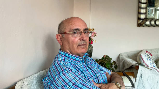 An old man, wearing eyeglasses, looks directly at the camera, his wise gaze filled with a lifetime of experiences, as he sits comfortably on a sofa in his home