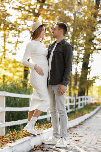 Pregnancy and Parenthoods concept - smiling man is walking with pregnant wife and looking to each other in autumn park. Happy styling young family is relaxing outdoors on weekend on sunny day