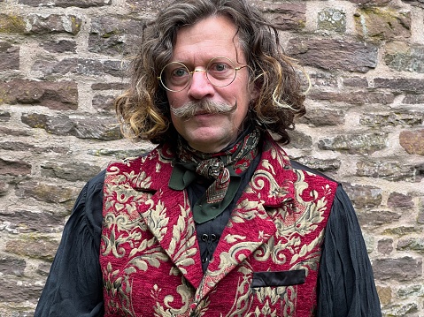 Middle aged man with long curly hair and a moustache. Wearing gold rimmed glasses and regency costume. Photo taken in the garden by a stone wall