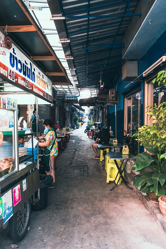 Thailand, Bangkok - February 9, 2023: Narrow , small street in Bangkok full of tiny tables where people sit eating their lunch. At the beginning of the street, there is a hand cart where a woman is serving fried chicken. A cheap place where locals are coming for lunch in Bangkok.