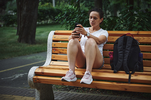Young multi-ethnic woman using mobile phone, checking new mobile app, scrolling news feed, sitting on a wooden bench in the city park. People. Mobile applications. Communication. Technology. Lifestyle