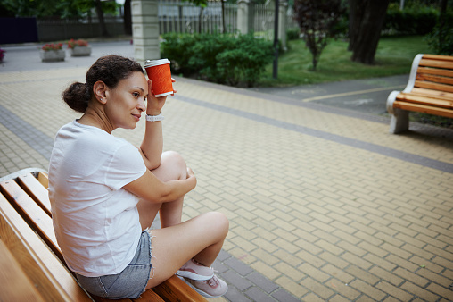 Beautiful multi-ethnic sporty serene pensive woman in blue jeans shorts, holding takeaway coffee in paper mug, sitting on wooden bench in the park. People. Lifestyle. Leisure activity. Recreation