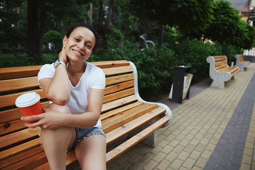 Attractive Latin American young female tourist smiling looking at camera, holding a cardboard mug with takeaway coffee, sitting on wooden bench in the summer park. People. Lifestyle. Leisure activity