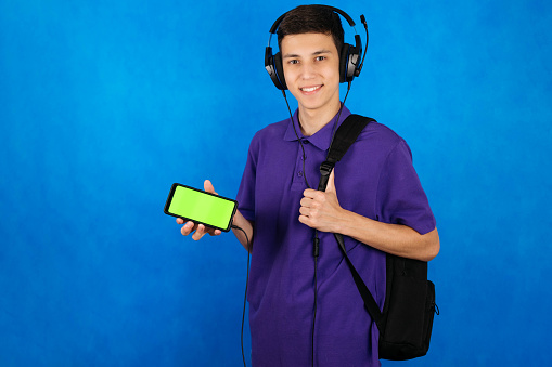 Close-up portrait of a cute teenager in headphones, with a backpack and a chroma key smartphone. Cope space.