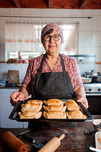 Flavors of Heritage: Latin Elderly Woman Preparing Chilean Baked Beef Empanadas in the Warmth of Her Home Kitchen. Looking at camera