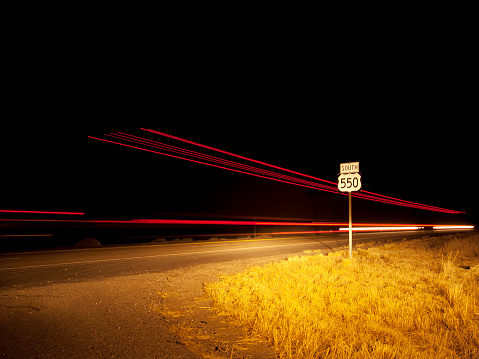 red and white light streaks pass a desert highway landscape road sign for highway 550 north on the shoulder of the road.  night shot with long exposure.  horizontal composition with copy space taken in northern new mexico.