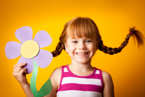 Whimsical, color photo of a happy, red-haired girl with upward braids holding a paper flower.