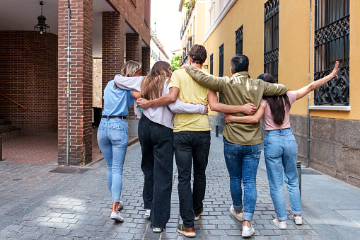A candid shot capturing the happiness and camaraderie of five friends walking arm in arm, their backs facing the camera, as they stroll along a bustling city street.