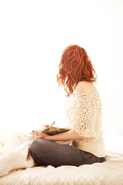 Beautiful woman with red hair sitting in bed writing in her journal backlit by light from a window.  Soft focus and washed out look intentional.