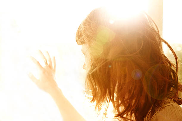 Beautiful woman with red hair backlit by the sun looking down with her hand on a glass window, nature scene beyond.  Washed out look, lens flare and soft focus intentional.